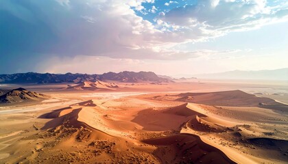 An aerial view of a vast desert landscape featuring undulating sand dunes casting long shadows, with a mountain range in the distance under a dynamic sky with c