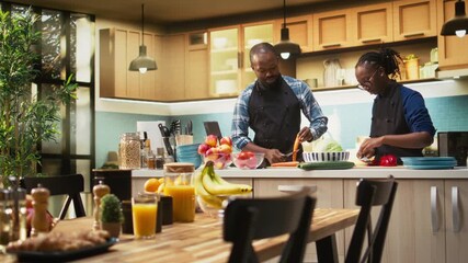 African American couple chopping fresh bell peppers and carrots for organic salad, preparing vegetarian food at home. Lovers in aprons cooking together and having fun in the kitchen. Camera A. - Powered by Adobe