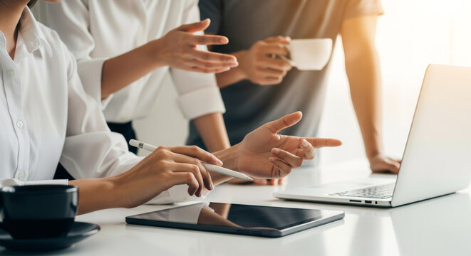 A diverse team collaborates around a white desk, using a laptop and tablet in a brightly lit office space during daytime. Teamwork, digital and internet concept.