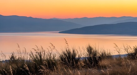 Golden hour panorama over a serene lake, framed by majestic mountains under a sky painted with soft orange and pink hues, viewed from a tranquil grassy hillside