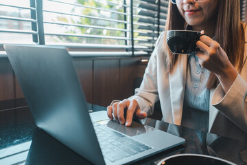 Businesswoman using a laptop and sipping coffee in a contemporary office with natural lighting.