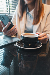 Professional woman in office attire holding a smartphone with a cup of coffee and laptop on a glossy table.