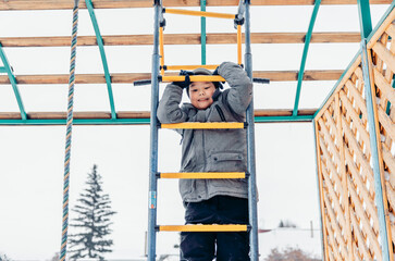 A young Asian boy climbs a yellow ladder in a snowy playground. Pine trees are visible in the background, creating a winter atmosphere, child safety on the street