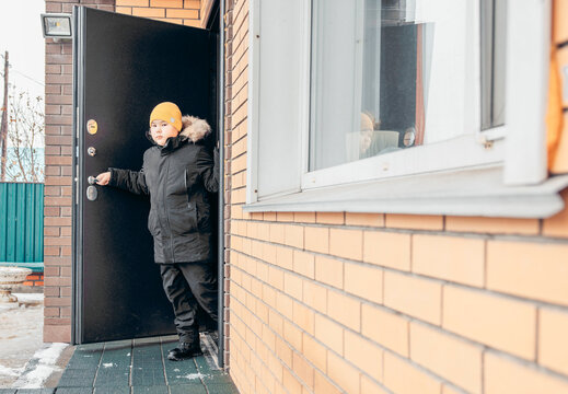 A young Asian boy with a yellow hat stands at the door of a brick house during winter. Snow is visible on the ground outside, child safety on the street