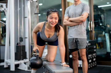 Pretty asian woman client holding kettlebell exercising on bench fitness trainer man standing in gym