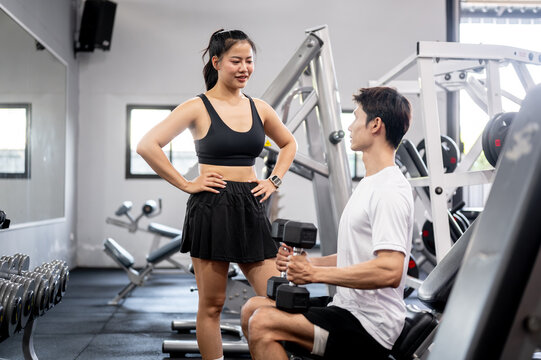 Asian man client holding dumbbell talking to pretty woman fitness trainer after exercising in gym. - Powered by Adobe