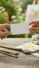 Hands exchanging mock up blank card, surrounded by art supplies and colorful background, evokes