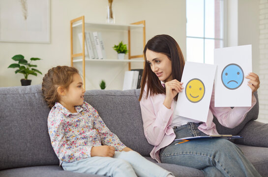 Mother showing cards with sad and happy emoji to smiling daughter to practice communication skills together at home. Psychologist using cards with different expression to teach preschool girl