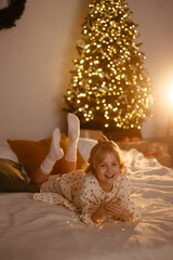Caucasian girl, about 9 years old, blonde, in white dress with small patterns, lying on bed holding string lights, Christmas tree glowing in background. Concept: childhood, holiday, family, joy.