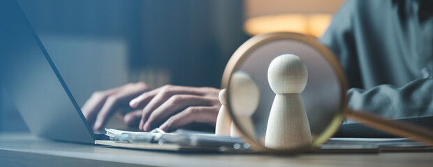 Obraz premium Magnifying glass focusing on wooden figures with a person typing on a laptop, representing recruitment, talent search, HR evaluation, and candidate selection processes.