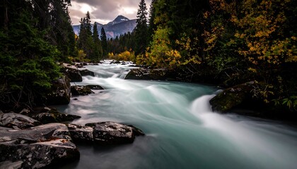 A fast-flowing river cuts through a wooded gorge, with autumn colors on the trees. Mountains rise in the distant background under a cloudy sky