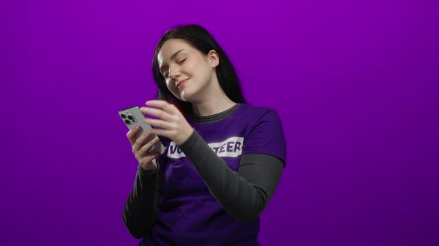Woman volunteer using smartphone in purple shirt against vibrant purple background showcasing a modern tech-savvy image capturing the essence of community service.