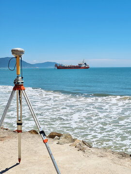 GNSS Global Navigation Satellite System receiver, topographic equipment for location surveys used in geodesy, engineering and coastal mapping work, and dredger ship at sea seen from the beach sand.