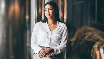 Smiling young businesswoman in white blouse standing near window with natural light