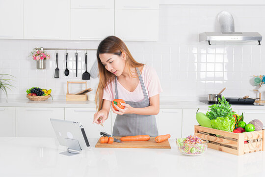 Attractive woman is using a digital tablet to follow a recipe while actively preparing fresh ingredient in like carrot and holding capsicum in a bright modern white kitchen.