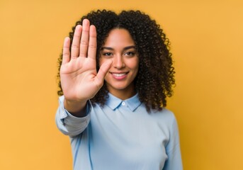 Young woman with curly hair raising her hand in a stop gesture, smiling confidently against a vibrant yellow background, conveying a message of prohibition or refusal