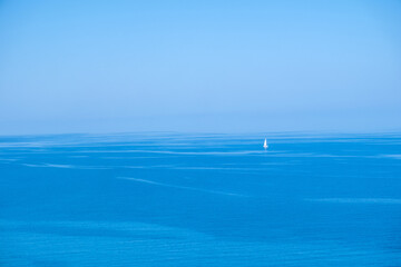 Lone white sailboat upon calm blue ocean midday, viewed from afar under clear bright sky
