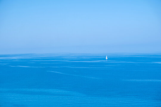 Lone white sailboat upon calm blue ocean midday, viewed from afar under clear bright sky
