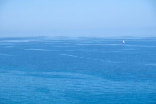 Solitary white sailboat upon calm blue sea horizon under clear daylight sky, minimal and serene