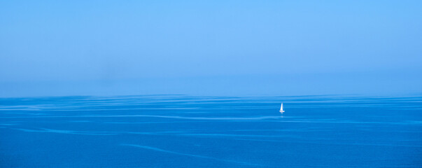Solitary white sailboat drifting upon calm blue sea under clear sky, evoking peaceful minimal mood