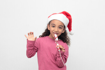 4-year-old Latina girl with curly brown hair wearing a santa hat sings with a candy cane to celebrate Christmas and New Year