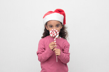 4-year-old Latina girl with curly brown hair wearing a santa hat sings with a candy cane to celebrate Christmas and New Year