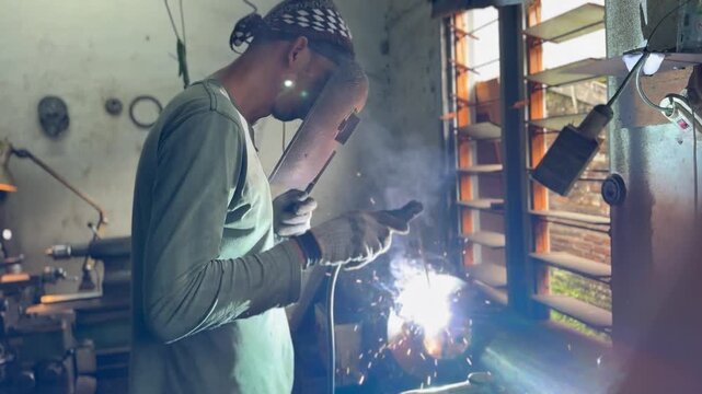 Craftsman welding metal in a workshop illuminated by a task lamp. The industrial cinematic video camera pans to the left. Great for technical vocational education videos.