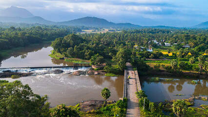 Fototapeta premium Aerial view of Attala Bridge and Cheramangalam check dam in Palakkad, Kerala, India, with river waters, lush green landscapes, coconut groves, and distant Western Ghats hills under a cloudy blue sky.