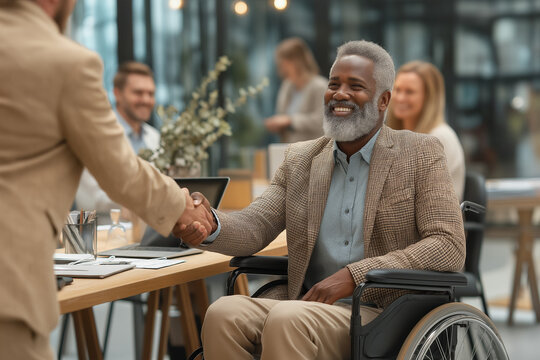 Smiling elderly black man in wheelchair shakes hands with colleague in modern office setting.