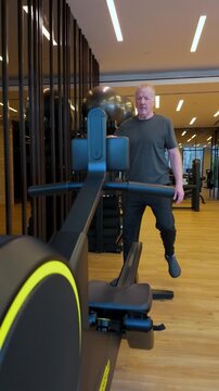 Vertical Shot Of A Senior Man Walking And Sitting On A Rowing Exercise Machine In A Indoor Gymasium.