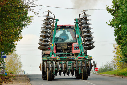 Tractor transporting large disc tillage equipment along tight rural road. Tractor with disc tillage takes up half narrow road, hard to pass, cars risk collision. Farm machinery danger, risks for cars