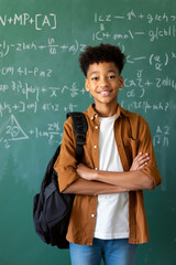A boy stands with arms crossed, wearing a brown shirt and backpack, in a classroom with a green chalkboard covered in complex math equations. Generative AI.