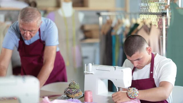 Young guy tailor sews on machine elderly man assistant drawing pattern on paper in workshop