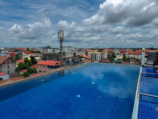 Tranquil rooftop infinity pool with deep blue water offers a luxurious, panoramic view over the urban landscape and diverse architecture of Siem Reap, Cambodia, under a dramatic and cloudy summer sky.