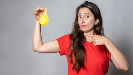 Woman in red T-shirt holds a yellow balloon while pointing at it in a simple studio setting