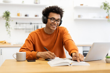 A young man wearing headphones and glasses smiles while taking notes and using a laptop at a wooden table.