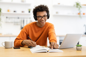 Obraz premium A young man wearing headphones smiles while studying at a table with a laptop and notebook.