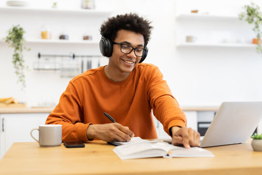 A young man wearing headphones and glasses smiles while taking notes and using a laptop at a wooden table.