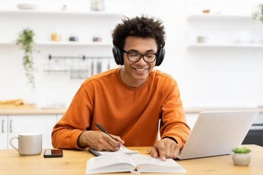 A young man wearing headphones and glasses smiles while studying with a laptop and taking notes in a notebook at home.