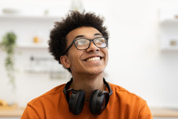 A young Black man with glasses smiles while looking up, with headphones around his neck.