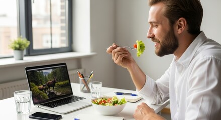 A smiling man enjoys a healthy salad for lunch while working at his laptop in a bright and modern office​