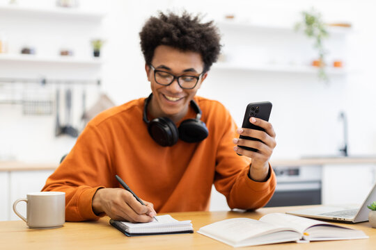 A young man with glasses smiles while looking at his phone and writing in a notebook at a wooden table. - Powered by Adobe