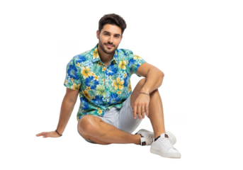 A stylish young man with dark hair and a beard wearing a vibrant floral print short sleeved shirt and white shorts sits relaxed isolated on transparent background