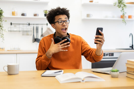 A young man with glasses is on a video call in his kitchen, looking surprised and gesturing with his hands. - Powered by Adobe