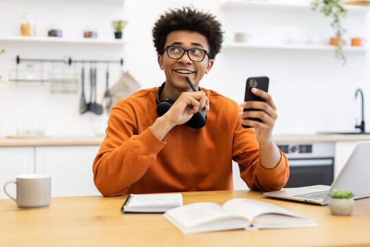 A young man with glasses is holding a phone and thinking while sitting at a table with a book and a laptop. - Powered by Adobe