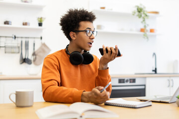 A young man with glasses is talking on his phone while sitting at a table in a modern kitchen.