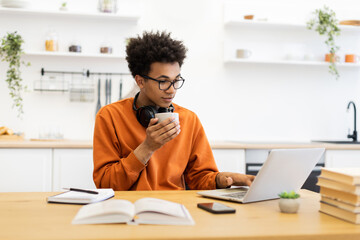 A young man with glasses is working on a laptop while drinking coffee at home.