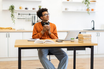 A young man with glasses enjoys a cup of coffee while working on his laptop in a bright, modern kitchen setting.