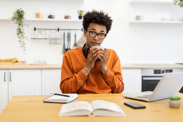 A young man with glasses enjoys a warm drink while working at a table in a modern kitchen setting.