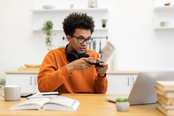 A young man with glasses and headphones is engrossed in a video game, holding a controller while sitting at a table with a laptop.